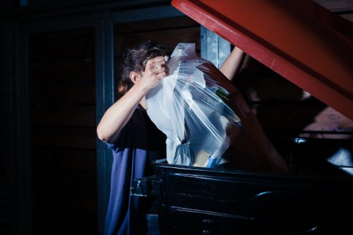 A person is seen from the side, leaning over a large black waste bin, lifting a clear plastic bag filled with assorted rubbish. The individual is wearing a dark t-shirt and is placing or removing waste materials into the bin, which is situated outdoors on a paved surface. In the background, there is a dark wooden structure, possibly a shed or garage, with a red metal roof visible at the top. The scene suggests a rubbish removal process, typical of household or property clearance work, consistent with services offered by Bermondsey House Clearance. The lighting indicates an overcast or shaded environment, emphasizing the plain, functional context of waste disposal activities in a residential or garden setting within Bermondsey, London.