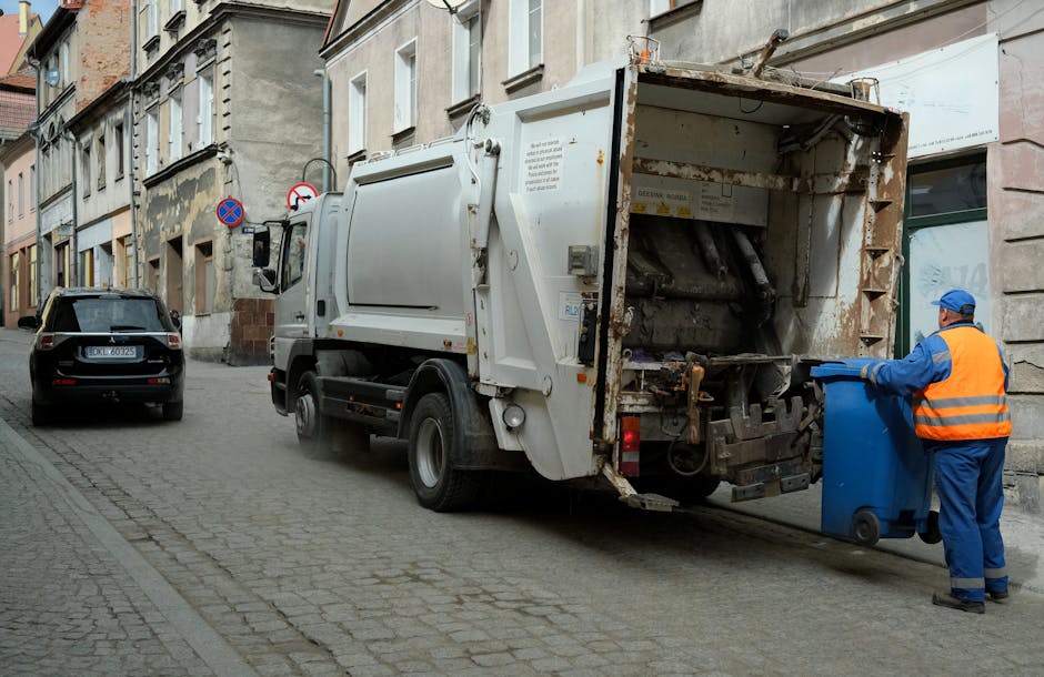 A rusted, red metal skip with a weathered surface and vertical ridges is positioned against a large, smooth, grey stone wall on a paved urban sidewalk. The skip is partially filled or weathered, with